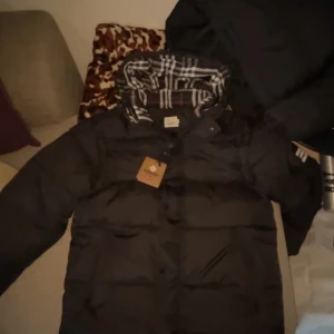 Bufberry jacka väst - Säljer en stilren svart dunjacka från Burberry med klassiskt rutigt foder. Jackan har knappar och en hög krage för extra värme. Perfekt för kyliga dagar.