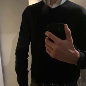 Polo Ralph Lauren Svart Tröja  - Snygg svart stickad tröja från Polo Ralph Lauren med lila broderad logga på bröstet. Tröjan har en v-ringad hals, långa ärmar och är i 100% pima cotton, perfekt för en stilren look. Passar M/L. Nypris ca 1500-2000 kr. Pris kan diskuteras.