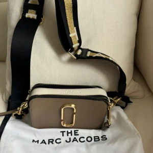 Beige och svart axelväska från Marc Jacobs - Snygg axelväska från Marc Jacobs i beige och svart med gulddetaljer. Väskan har en bred, justerbar axelrem med svarta och guldiga ränder. Modellen är snapshot