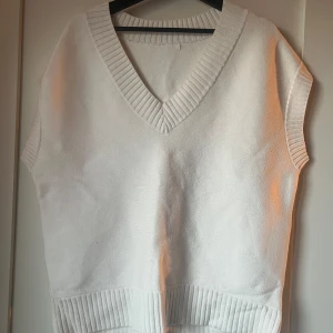Vit stickad väst - Säljer en stilren vit stickad väst med v-ringad design och ribbade kanter. Perfekt för lager-på-lager-stil och passar till många olika outfits.
