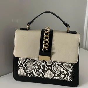 Stilren handväska med ormmönster - Snygg handväska i beige och svart med ormmönstrad detalj. Väskan har en elegant kedja och ett praktiskt handtag. Perfekt för att ge din outfit en trendig touch.