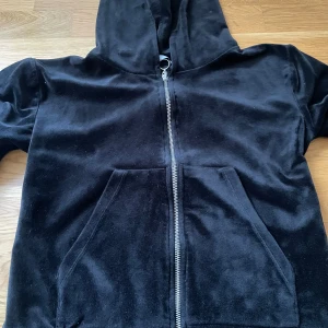 BUDA PRIS: Svart velour hoodie - Snygg svart hoodie i mjuk velour med dragkedja framtill och praktisk känguruficka. Perfekt för en avslappnad stil med en touch av lyx. Den har långa ärmar och en bekväm huva.