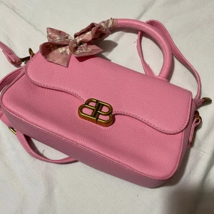 Rosa axelväska med rosett - Snygg rosa axelväska i syntetmaterial med ett elegant guldfärgat spänne. Väskan har en söt rosett knuten runt handtaget och en justerbar axelrem. Perfekt för att ge en färgklick till din outfit!