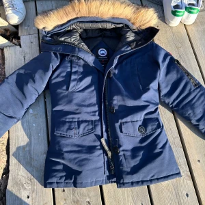 Blå dunjacka med päls från Canada Goose - Säljer en blå dunjacka från Canada Goose med avtagbar päls på huvan. Jackan har dragkedja och knappar framtill samt flera praktiska fickor. Perfekt för kalla vinterdagar. Har ingen aning om den är äkta eller fejk. Noll koll på de 