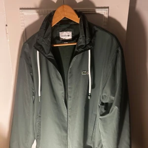 Grön vindjacka från Lacoste - Säljer en stilren grön vindjacka från Lacoste med dragkedja och justerbar huva. Jackan är tillverkad i återvunnen polyester och är vattenavvisande, perfekt för blåsiga dagar. Den har en broderad logotyp på bröstet och praktiska fickor. Köpt i somras andvänt 2 gånger som ny