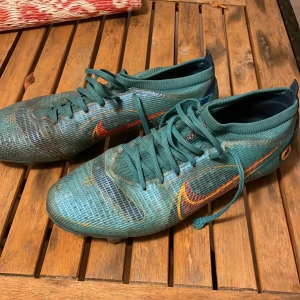 Blåa fotbollskor från Nike - Säljer ett par gröna Nike fotbollsskor med snörning och orange detaljer. Skorna har en syntetisk ovandel och är designade för optimal prestanda på planen.