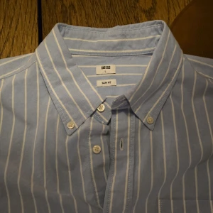 Blå och vit randig skjorta från Uniqlo - Snygg blå och vit randig skjorta från Uniqlo i slim fit. Skjortan har en klassisk krage och knappar framtill samt en bröstficka. Perfekt för en stilren look!