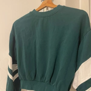 Grön sweatshirt med vita ränder - Säljer en grön sweatshirt med vita ränder på ärmarna. Tröjan har en ribbad kant vid midjan och ärmarna, den är använd ett fåtal gånger men är k bra skick. 