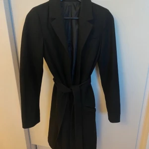 Svart kappa med bälte - Elegant svart kappa med bälte och långa ärmar. Kappan är frpn ginatricot och har knappar längst med! Sitter vid knäskålen på mig som är 160cm!Köpt på Vinted men säljer vidare