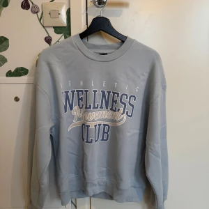 Ljusblå sweatshirt från Gina Tricot - Säljer en ljusblå sweatshirt från Gina Tricot med texten 'Athletic Wellness Movement Club' i vitt och blått. Tröjan har en rund hals och långa ärmar, perfekt för en avslappnad stil.