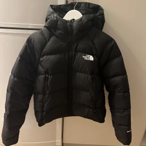Svart dunjacka från The North Face - Säljer en svart dunjacka från The North Face med huva och dragkedja. Jackan är perfekt för kyliga dagar och har en klassisk pufferdesign. Den är långärmad och har en liten logga på bröstet. kan diskutera pris