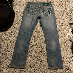 Blå jeans från True Religion - Säljer ett par blå jeans från True Religion i modellen Ricky Relaxed Straight. De har snygga detaljer på bakfickorna med gröna och vita sömmar. Perfekta för en avslappnad stil.