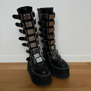 DAMNED-318  ’Svarta platåskor med spännen’ - Snygga svarta platåstövlar i veganskt läder. Med flera justerbara spännen och en hög platåsula. Perfekta för att ge en edgy look till din outfit. Stövlarna har en dragkedja på baksidan för enkel på- och avtagning. (Orginal pris 2000kr)