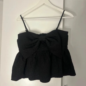 Svart topp med rosett - Snygg svart topp med smala axelband och en stor dekorativ rosett framtill. Perfekt för en kväll ute eller en speciell tillställning. Toppen har en elegant och stilren design.