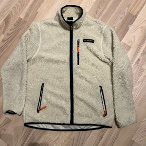 Beige fleecejacka från Ocean Works - Säljer en beige fleecejacka från Ocean Works med dragkedja och praktiska fickor. Jackan har en broderad logga på bröstet och kontrasterande svarta detaljer. Perfekt för kyliga dagar! Passar M