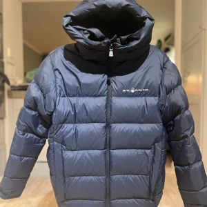 Sail Racing cloud down hood  - Säljer en mörkblå dunjacka från SailRacing med hög krage och huva. Jackan har en dragkedja framtill och är tillverkad av högkvalitativt dun med 600 fill power. Passar strl S. Perfekt för kalla dagar. Använd sparsamt under 1 säsong.