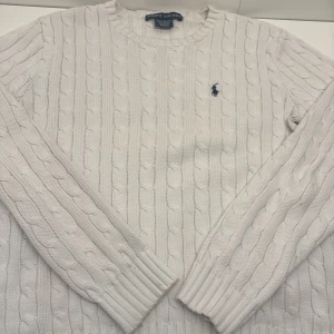 Vit kabelstickad tröja från Ralph Lauren - Säljer en stilren vit kabelstickad tröja från Ralph Lauren med deras ikoniska logga broderad på bröstet. Tröjan har långa ärmar och ribbade muddar vid ärmslut och nederkant. Perfekt för en klassisk look! Skriv om intresserad!!
