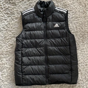 Svart dunväst från Adidas - Snygg svart dunväst från Adidas med dragkedja och klassiska vita ränder på axlarna. Perfekt för kyligare dagar och sportiga aktiviteter. Västen har en hög krage och är quiltad för extra värme.