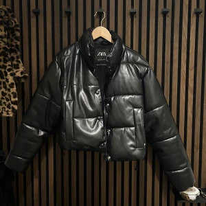 Svart pufferjacka från Zara - Snygg svart pufferjacka från Zara i syntetmaterial. Jackan har en quiltad design med knappar och dragkedja. Perfekt för kyligare dagar.
