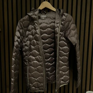 Brun pufferjacka från Strova - Snygg brun pufferjacka från Strova med dragkedja och huva. Perfekt för kyliga dagar med sin quiltade design som håller värmen. Jackan har en modern look och passar bra till vardags.