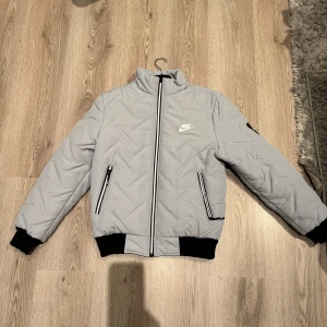 Grå Nike jacka  - Snygg grå jacka med märket Nike och svarta detaljer. Jackan har dragkedja framtill och två fickor med dragkedjor. Ribbstickade muddar vid ärmslut och nederkant. Storlek L. (Köparen står för frakten) 