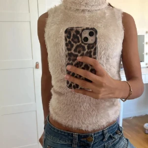 Fluffig topp - Säljer en fluffig beige ärmlös topp med hög krage. Perfekt för en trendig och mysig look. Passar bra till jeans eller kjol för en stilren outfit.