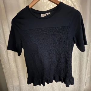 Svart topp med volangdetalj - Snygg blå topp med korta ärmar och volangdetalj vid nederkanten. Toppen har en smockad textur på framsidan som ger en fin passform. 