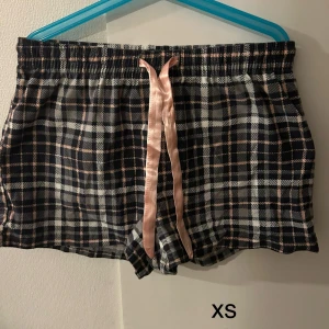 Rutig pyjamasshorts med rosa knytband - Mysiga rutig pyjamasshorts i svart, vit och rosa med ett sött rosa knytband i midjan. Perfekta för en avslappnad kväll hemma. Elastisk midja för extra komfort.