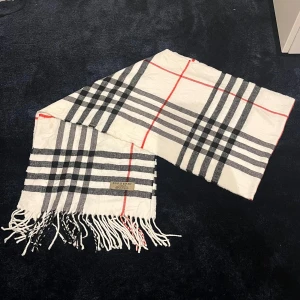 Rutig halsduk från Burberry - Snygg rutig halsduk från Burberry i klassiskt mönster med svarta, grå och röda ränder. Tillverkad i mjukt material med fransar i ändarna. Perfekt för att hålla stilen i kallare väder.