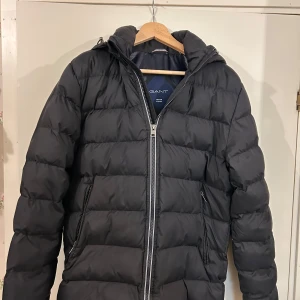 Svart dunjacka från GANT - Säljer en stilren svart dunjacka från GANT med huva och dragkedja. Jackan är quiltad och har långa ärmar, perfekt för kyligare väder. Säljer för den är för liten för mig nu, men är as snygg. Köpt för ungefär 3000 kr och ser helt ny ut 