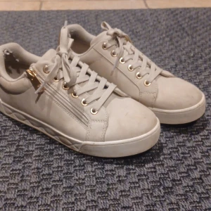 Beige sneakers med gulddetaljer - Snygga beige sneakers med gulddetaljer och snörning. Skorna har en dragkedja på sidan och en quiltad sula för extra stil. Perfekta för en trendig look. 