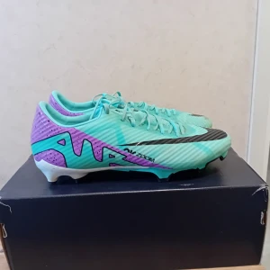 Turkosa och lila fotbollsskor - Snygga fotbollsskor i turkos och lila med svarta detaljer. Skorna har snörning och en slitstark sula med dobbar för bra grepp på planen. Perfekta för fotbollsträning och matcher.