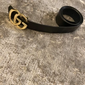 Svart skärp med guldspänne - Snyggt svart skärp med ett iögonfallande guldspänne i form av dubbla G:n. Perfekt för att ge en lyxig touch till din outfit. Skärpet är tillverkat i skinn och har en klassisk design som passar till många olika stilar.