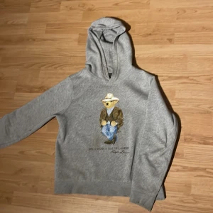 Grå hoodie från Ralph Lauren (14-16) - Säljer en stilren grå hoodie från Ralph Lauren med en cool björntryck på framsidan. Tröjan har långa ärmar och en klassisk huva. Perfekt för en avslappnad look. Skick 8/10 storlek 158 (14-16)