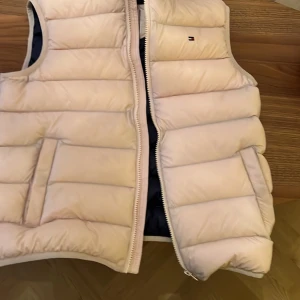 Rosa dunväst från Tommy Hilfiger - Snygg rosa dunväst från Tommy Hilfiger med dragkedja framtill. Perfekt för kyligare dagar och ger en stilren look. Västen har en diskret logga på bröstet och är vadderad för extra värme.