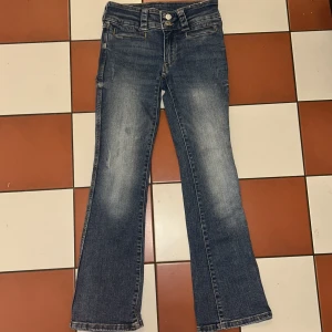 Blå bootcut jeans från H&M - Snygga blå bootcut jeans från H&M med låg midja och justerbar midja. Perfekta för en avslappnad stil med klassisk femficksdesign och dubbla knappar i midjan.