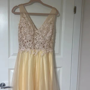 Champagnefärgad långklänning med spetsdetaljer - Elegant långklänning i champagnefärg med v-ringad spetsöverdel och blommönster. Kjolen är i ett lätt och flödande material som ger en vacker siluett.