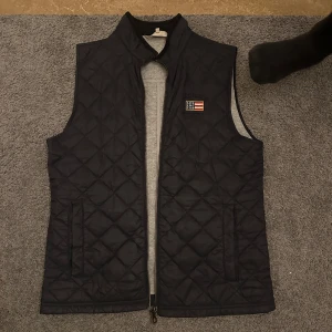 Svart quiltad väst från Lexington - Snygg svart quiltad väst från Lexington med dragkedja och ett amerikanskt flaggmärke på bröstet. Perfekt för lager-på-lager under kyliga dagar. Säljs också pågrund av att den är lite för stor på mig