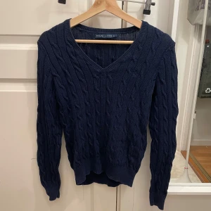 Mörkblå stickad tröja från Ralph Lauren - Säljer en snygg mörkblå stickad tröja från Ralph Lauren med kabelstickat mönster. Tröjan har en v-ringad hals och långa ärmar, perfekt för kyligare dagar. Ett klassiskt plagg som passar till många tillfällen 💕