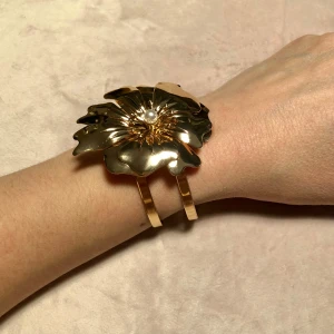 Stort guld armband med pärla och blomma  - Fint armband jag fått i julklapp men tycker de är lite för stort för min smak. De är en stor asymmetrisk blomma med en pärla i mitten. Rostfritt stål tror jag eller mässing med guld utan på. Skall inte tvättas utan putsas med duk. Helt nytt och oanvänt och justerbart 