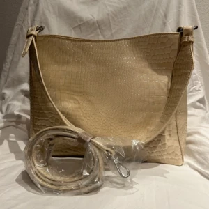 Beige axelväska från HVISK - Ordinarie pris för över 1000 kr! Snygg beige väska från danska märket HVISK, syntet med krokodilmönster. Väskan har en justerbar axelrem och flera fack med dragkedja inuti för praktisk förvaring. Höjd: 25,5 cm & Bredd: 31 cm. Använd endast en gång & har topp skick!