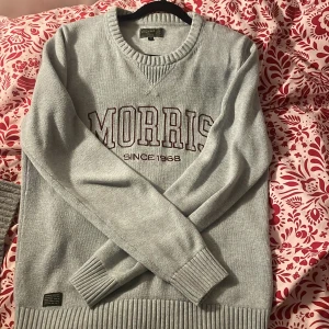 Grå tröja från Morris - Säljer en stilren grå tröja från Morris med texten 'Morris Since 1968' på bröstet. Tröjan har en rund halsringning och ribbade muddar vid ärmslut och nederkant. Perfekt för en avslappnad look.