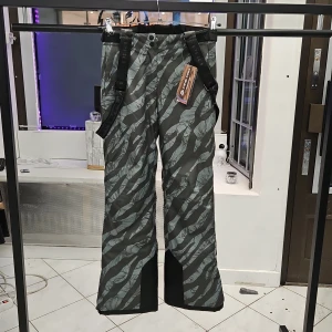 Gröna mönstrade byxor från Alpine Pro - Snygga gröna byxor från Alpine Pro med ett unikt mönster. De har justerbara hängslen och knappar i midjan för extra komfort. Perfekta för utomhusaktiviteter. Storlek XS, nya med etikett.