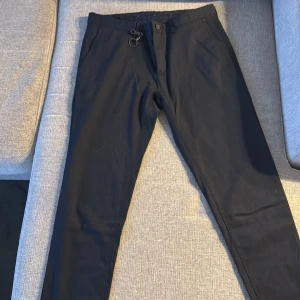 Svarta kostymbyxor med knappdetaljer - Snygga svarta kostymbyxor med stilrena knappdetaljer bak och en praktisk nyckelring framtill. Byxorna har en klassisk passform och är perfekta för en dressad look.