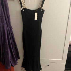 Svart långklänning med volangdetaljer - Elegant svart långklänning med volangdetaljer i nederkant och kontrasterande axelband. Klänningen har en tight passform och är perfekt för festliga tillfällen. Aldrig använt utan endast provat. Köptes på plick och säljer pga att den sitter lite tajt. Storlek XS. 