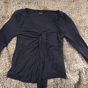 Mörkblå topp från Gina Tricot - Säljer en mörkblå topp från Gina Tricot i storlek XS. Toppen har en v-ringad design med knytning framtill och långa ärmar. Perfekt för en stilren look. Är dock lite nopprig. 
