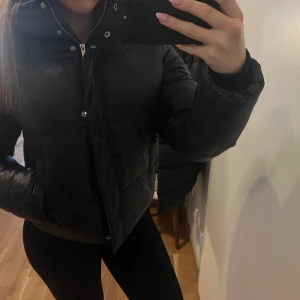 Svart pufferjacka - Säljer en svart pufferjacka med dragkedja och knappar. Jackan har en huva och är quiltad för extra värme. Perfekt för kyliga dagar.