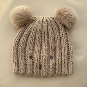 Beige stickad mössa med pom-poms - Söt beige stickad mössa med två stora pom-poms på toppen. Mössan har ett ribbat mönster och små broderade detaljer framtill. Perfekt för kyliga dagar!