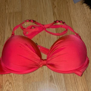 Röd bikiniöverdel med gulddetaljer! - Snygg röd/orange bikiniöverdel med vadderade kupor och knytband i nacken. Den har guldfärgade detaljer på axelbanden som ger en lyxig touch. Perfekt för en dag på stranden eller vid poolen. Aldrig använd.