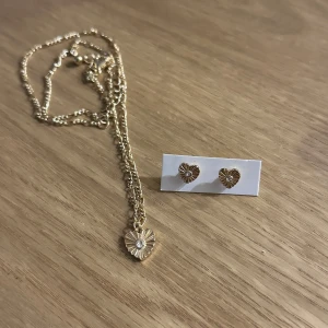 Guldhalsband och örhängen med hjärtdetaljer - Elegant set med ett guldhalsband och matchande örhängen. Halsbandet har ett hängsmycke i form av ett hjärta med en glittrande sten i mitten. Örhängena är också hjärtformade och kompletterar halsbandet perfekt. Oanvända!